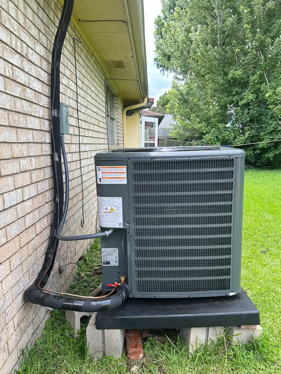 Newly installed gray outdoor AC condenser on a black pad beside a brick wall at a Kenner, Louisiana home, with black insulated line set running up the wall