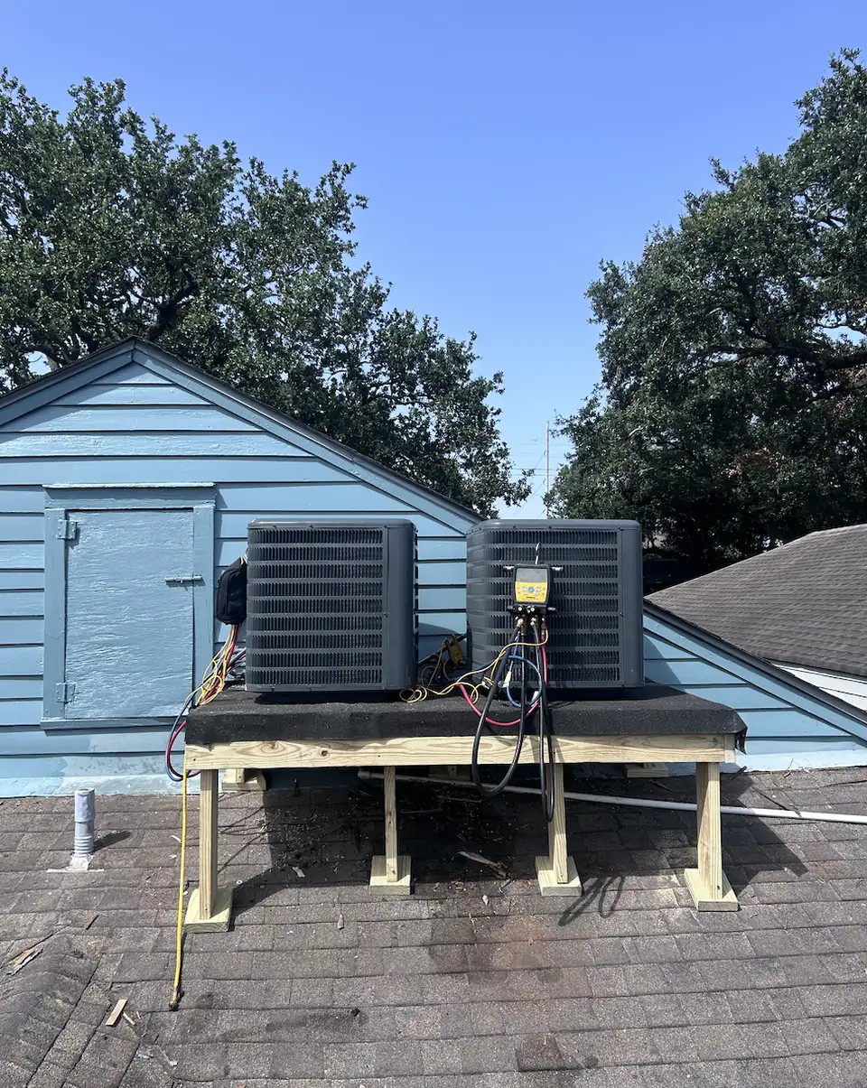 Two gray AC condensers set on a wooden rooftop platform above shingles at a raised Kenner, Louisiana home, with trees in the background